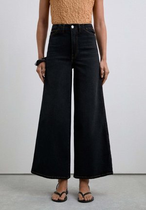 High-waisted black denim culottes with wide legs, featuring a front button closure and yellow stitching, worn with black flip-flops.
