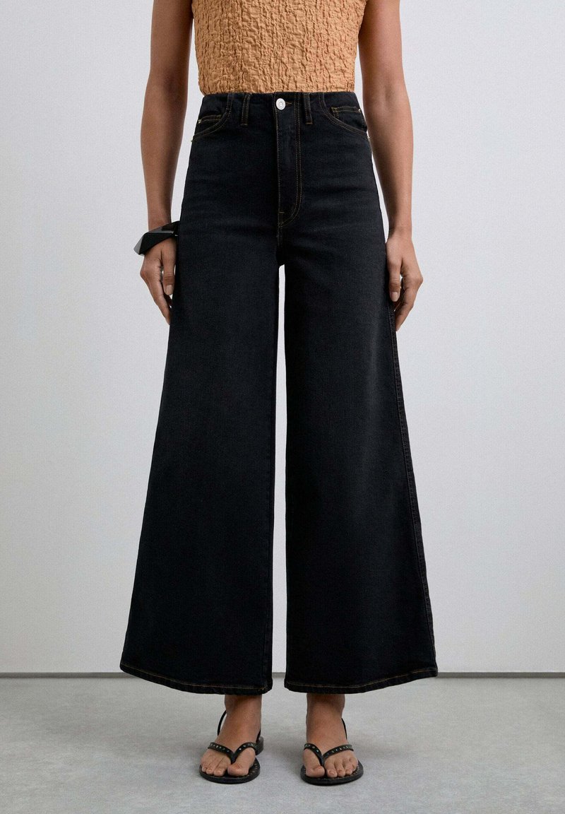 Hoch taillierte schwarze Denim-Culottes mit weitem Bein, ausgestattet mit einem vorderen Knopfverschluss und gelben Nähten, getragen mit schwarzen Flip-Flops.