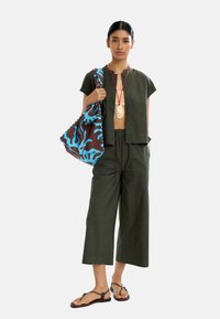 Zielona, krótka bluzka z guzikami, dopasowane szerokie spodnie, brązowa torba z niebieskim koralikowym wzorem i beżowe płaskie sandaly.
