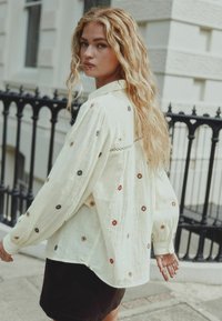 Cream-colored blouse with multicolored embroidered patterns. Long, loose sleeves and a buttoned collar. Soft, textured fabric.