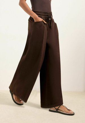 Person iført løstsiddende mørkebrune bukser med lommer og sorte sandaler, stående mod en ensartet, lys baggrund.