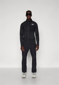 Črno jakno z zadrgo in hlače iz sintetičnega blaga, z žepom na prsih, nošene z sivimi pohodnimi čevlji in velikimi sončnimi očali.