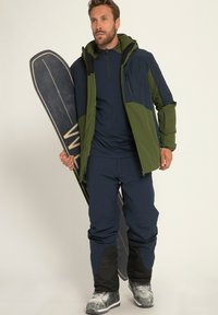 Skidragt med en grøn og marineblå jakke, marineblå baselag og blå skibukser med sorte detaljer. Personen holder et sort snowboard.