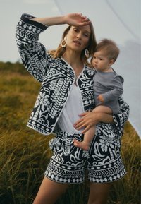 Schwarz-weiß gemusterte Jacke und Shorts-Set mit floralen und geometrischen Designs. Modell, das ein Kind in einem grauen Outfit vor einer grasbewachsenen Kulisse hält.