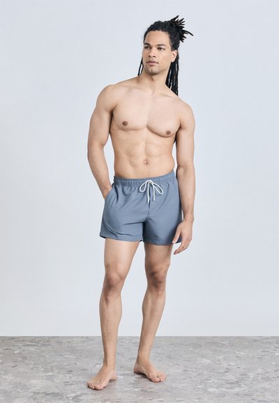 Άνδρας με γυμνό στήθος και κότσους στα μαλλιά, ντυμένος με ανοιχτό μπλε μαγιό, στέκεται ξυπόλητος σε δαπέδο με στίγματα, μπροστά σε απλό φόντο.