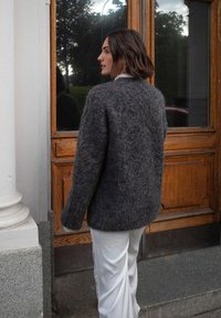 Pilkas, pūkuotas megztinis su laisvu kirpimu ir nuleistomis petnešomis, derinamas su baltomis kelnėmis, matomas iš nugaros prie medinių durų.