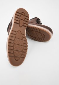 Hnedé kožené čižmy s textúrovanými gumovými podrážkami. Podrážky majú hlboké drážky a vzorovaný dizajn pre lepšiu trakciu. Viditeľné sú detaily šnúrok a šitia.