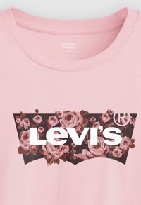 T-shirt en coton rose clair avec le logo "Levi's" blanc sur un motif floral avec de grandes roses sur un fond sombre, col rond, texture lisse.