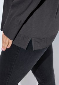 Maglione a maglia grigio scuro con accenti texturizzati all'orlo e spacchi laterali, indossato sopra jeans in denim neri. Dettaglio ravvicinato del capo.