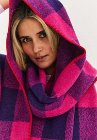 Wełniany szal w kratę w kolorach fuksji i purpury, o miękkiej fakturze i kontrastującym, czerwonym obszyciu na brzegach.