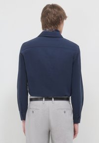 Camicia a manica lunga blu navy con una texture liscia, caratterizzata da un colletto classico e polsini abbottonati, abbinata a pantaloni grigio chiaro.