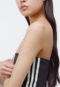 Čierna bezramienková súčasť oblečenia s bielymi vertikálnymi pruhmi; hladká látka; detailný pohľad na plece a ruku, vlasy voľne padajúce po boku.