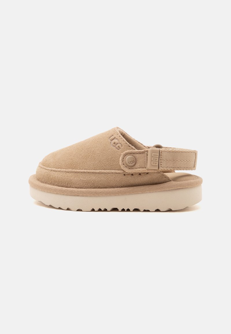 Μπεζ σουέντ παπούτσι slip-on με ανοιχτή φτέρνα, ρυθμιζόμενο λουράκι, ανυψωμένη σόλα και ανάγλυφο λογότυπο UGG στο πλάι και στο κουμπί του λουριού.