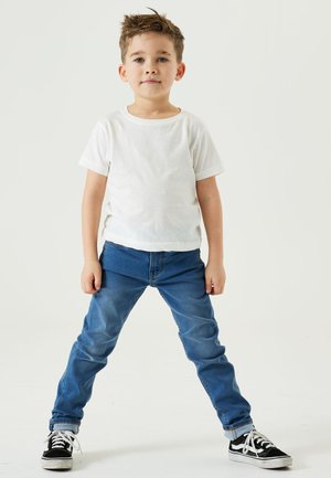 Witte t-shirt met korte mouwen gecombineerd met blauwe denim jeans. De jeans hebben een slank ontwerp met een licht vervaagde afwerking en opgerolde mouwen. Zwarte sneakers.