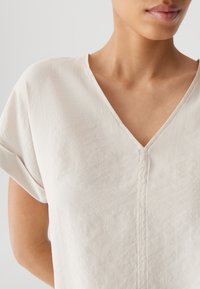 Helles beige Bluse mit kurzen Ärmeln und V-Ausschnitt, mit subtiler Textur und einer mittigen Nahtdetails. Glattes Material mit lockerer Passform.
