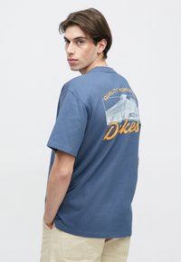 Νέος άνδρας με κοντά καστανά μαλλιά που φοράει μπλε γραφικό T-shirt και μπεζ παντελόνι, κοιτάζοντας πάνω από τον ώμο σε απλό λευκό φόντο.