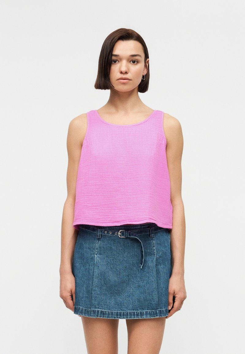 Femme aux cheveux courts et foncés portant un haut sans manches rose texturé et une mini jupe en jean avec ceinture, debout devant un arrière-plan uni.