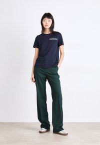 T-shirt en coton bleu marine avec texte imprimé ; associé à un pantalon large vert ; le mannequin se tient devant un fond uni, les mains dans les poches.