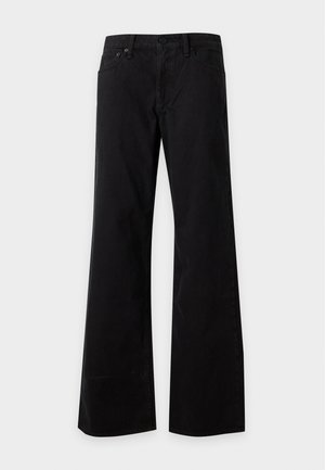 Femme debout, bras croisés, portant un pull gris foncé à large col, un pantalon noir à jambes larges et des chaussures noires sur un fond uni.