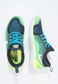 Běžecké boty Nike s prodyšným černým síťovaným svrškem, živými zelenými a modrými akcenty a flexibilní bílou podrážkou. Obsahují poutko pro snadné obutí.