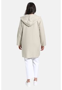Veste longue beige à capuche avec une texture douce, présentant une coupe décontractée et des poches latérales, portée sur un pantalon large blanc et des baskets.