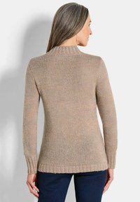 Beiger Strickpullover mit einem gerippten Kragen und Bündchen, der ein strukturiertes Muster und einen länger geschnittenen Rückensaum aufweist, kombiniert mit dunklen Hosen.