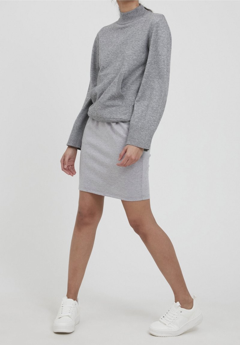 Personne portant un pull gris à manches longues et une jupe assortie avec des baskets blanches, debout devant un fond clair uni.