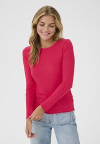 Long-sleeve top in bright pink ribbed fabric with a round neckline and scalloped hem, paired with light wash denim.