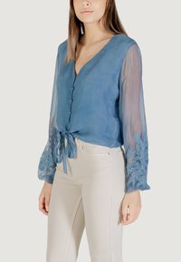 Blusa azul claro con escote en V, diseño con lazo en el frente y mangas transparentes que presentan patrones florales bordados y acentos de cuentas.
