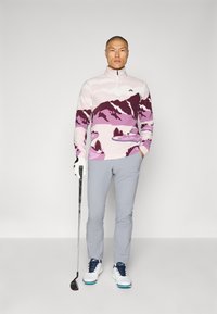 Pullover leggero rosa con chiusura lampo e motivo di montagne e alberi, abbinato a pantaloni grigi chiari e scarpe da golf bianche con dettagli blu. Bastone da golf tenuto nella mano sinistra.