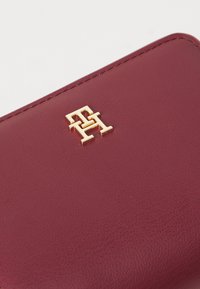 Bordeauxfarbene Lederbörse mit strukturiertem Oberflächenfinish und Hardware mit goldfarbener Logo-Prägung. Die Nähte sind entlang der Kanten sichtbar.