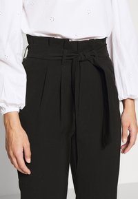 Svarta högmidjade byxor med veck och ett tygbälte, med en smidig textur. Kombineras med en vit långärmad skjorta med broderi.