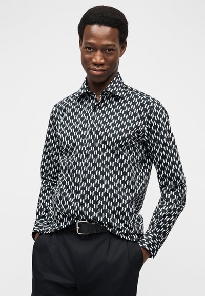 Homme portant une chemise boutonnée à motifs noir et blanc avec un pantalon noir et les mains dans les poches, sur fond uni.
