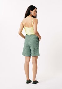 Groen gestreepte shorts met zijzakken, gecombineerd met een gele spaghetti-top. Model staat met de rug naar de camera, met groene schoenen aan.