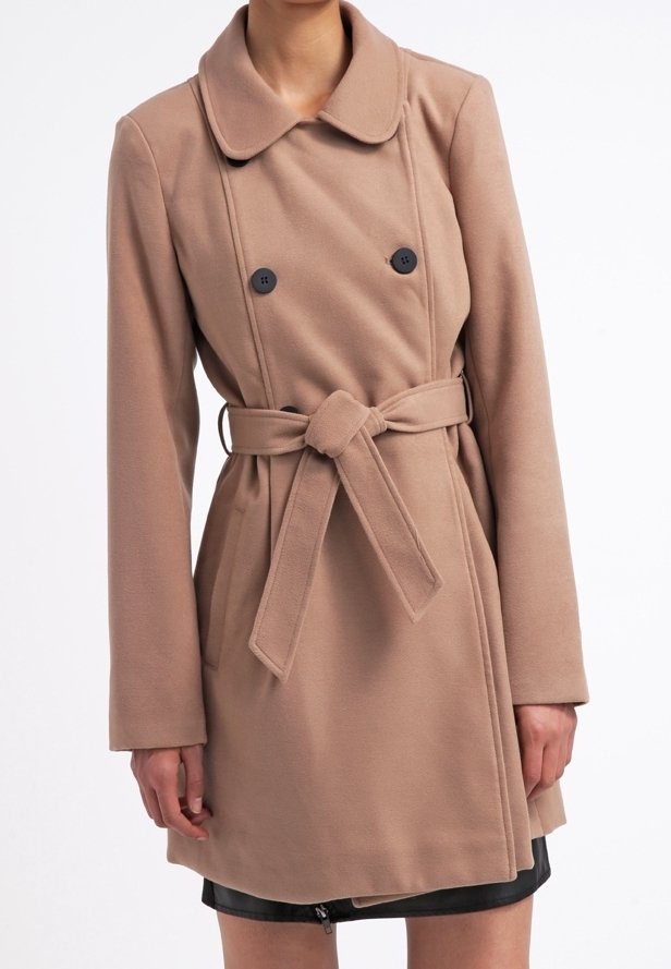 Kvinde iført en knælang, dobbeltbrystet kamelfarvet frakke med en bundet taljebælte over en sort nederdel.