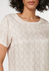 Lichtbeige korte mouw top met een subtiele logoprint, ronde halslijn en een gladde textuur. Geaccentueerd met een iets doorzichtig materiaal.