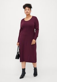 Woman standing wearing a long-sleeve burgundy midi dress, black ankle boots, holding a small black handbag against a white background.