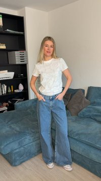 Γυναίκα με μακριά ξανθά μαλλιά στέκεται με τα χέρια στις τσέπες φορώντας λευκό μπλουζάκι με σχέδια, τζιν παντελόνι φαρδιάς γραμμής και λευκά αθλητικά παπούτσια σε σαλόνι.