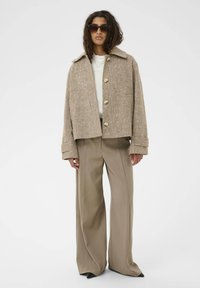 Rjava teksturirana skrajšana jakna z velikimi gumbi, v kombinaciji z visokimi bež hlačami in konicami črnimi čevlji. Model nosi sončna očala.