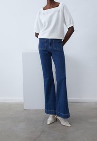 Vit topp med fyrkantig halsringning och korta puffärmar, i kombination med mörkblå utsvängda jeans. Modellen bär vita nätspetsiga klackar.