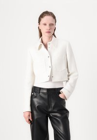 Veste blanche texturée cropped avec un devant à boutons, deux poches poitrine et un col classique, associée à un pantalon en cuir noir.