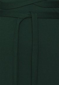 Ciemnozielony materiał o gładkiej fakturze, z wzorem z narzutką i akcentowanej talii związanej węzłem. Kolor jest głęboki i jednolity.