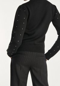 Schwarzer Strickpullover mit puffigen Ärmeln, verziert mit kleinen silbernen Akzenten. Kombiniert mit schwarzen Streifenhosen, die über Rückentaschen verfügen.