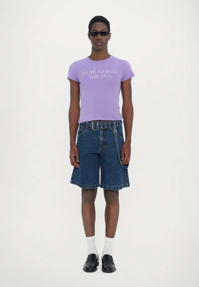 Tricou purpuriu ajustat cu textul "AICI VIN DISTRACTIA", combinat cu pantaloni scurți din denim, curea neagră cu detalii metalice, șosete albe și mocasini negri.