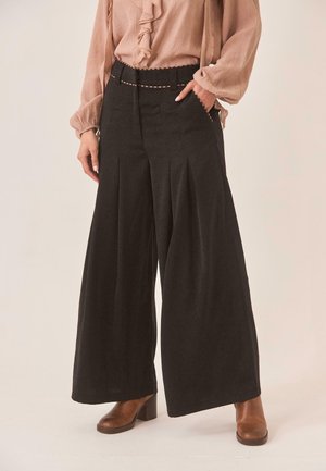 Mujer con pantalones negros de cintura alta y piernas anchas con pliegues, una blusa beige con volantes metida por dentro, y botines marrones de tacón.