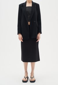 Top corto negro debajo de un blazer negro con un solo botón, combinado con una falda midi negra y chanclas negras. Textura de tela suave.
