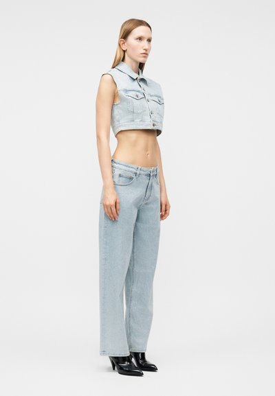 Γυναίκα στέκεται φορώντας κοντό αμάνικο τζιν μπουφάν σε ανοιχτό μπλε και τζιν παντελόνι με φαρδιά πόδια σετ, με μαύρες μπότες με τακούνι και μυτερή μύτη, σε λευκό φόντο.