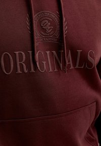 Gros plan d’un sweat à capuche bordeaux avec le texte en relief « ORIGINALS » et un emblème circulaire portant les mentions « Jack & Jones » et « Creative Dept. » au-dessus de la poche.