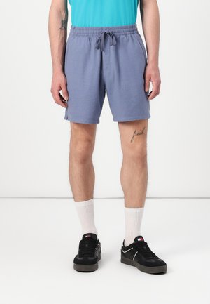 Men's athletic shorts in light blue fabric, elastic waistband with drawstring, side pockets, worn with white crew socks and black sneakers.