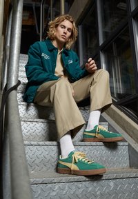 Baskets Puma vertes et jaunes avec une semelle marron texturée, portées avec un pantalon beige et une veste en velours côtelé bleu sarcelle, sur des escaliers métalliques.